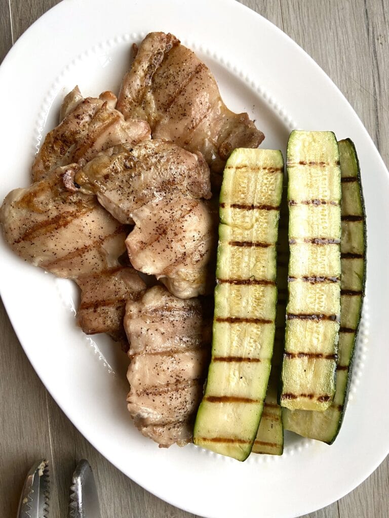 Top down shot of grilled boneless skinless chicken thighs and grilled zucchini on a white oval platter.