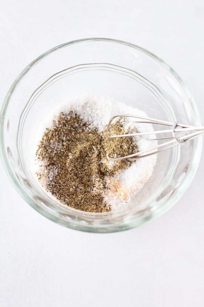 Close up of a small whisk stirring ground black pepper, kosher salt, and garlic powder together in a small clear bowl that rests on a white surface.