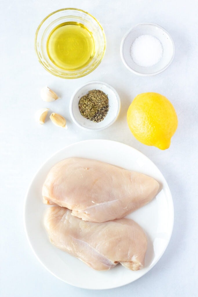 Top down shots of ingredients for grilled lemon chicken, including a white plate with two chicken breasts on it, a lemon, bowls with salt, herbs, and oil, and some garlic cloves.