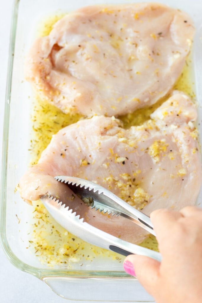 Tongs flipping over raw chicken breasts resting in a glass dish with a lemon marinade on them.