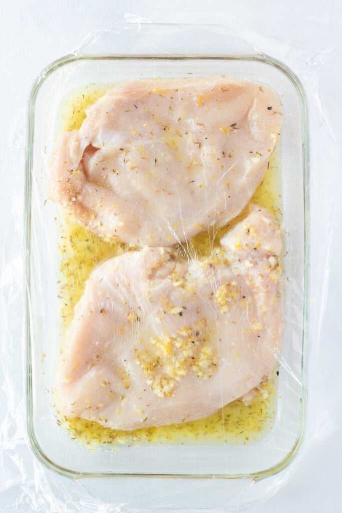 Top down shot of two raw chicken breasts in a flat glass dish in a lemon herb marinade with plastic wrap covering the dish.
