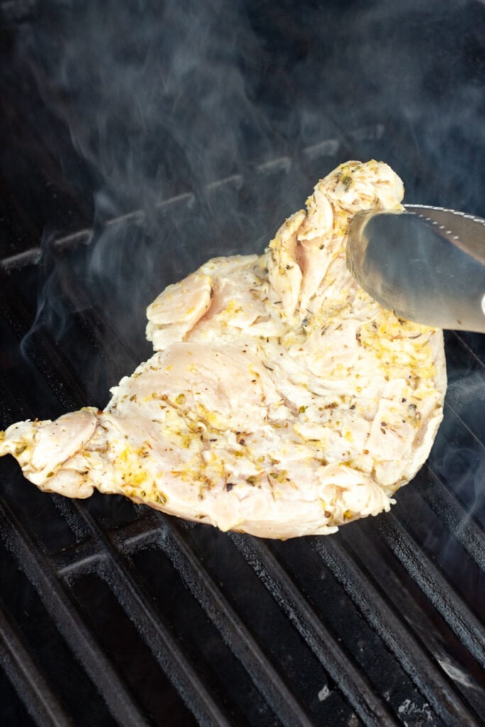 A pair of tongs placing a lemon marinated chicken breast on a hot grill.