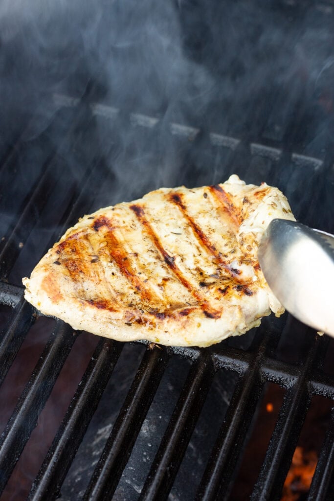 A pair of metal tongs flipping a chicken breast over on a grill.