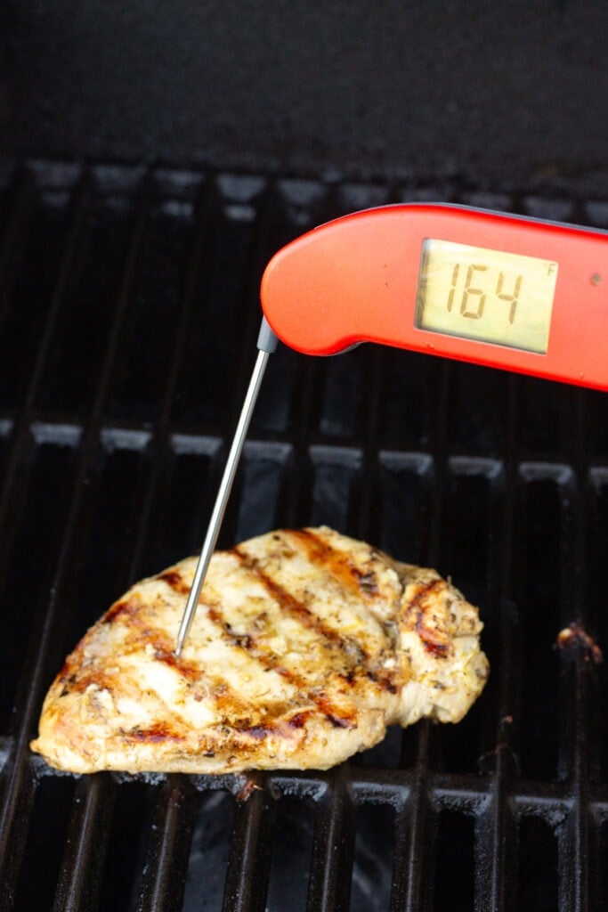 A red meat thermometer measuring the internal temp of a cooked chicken breast on a grill at 164 degrees F.