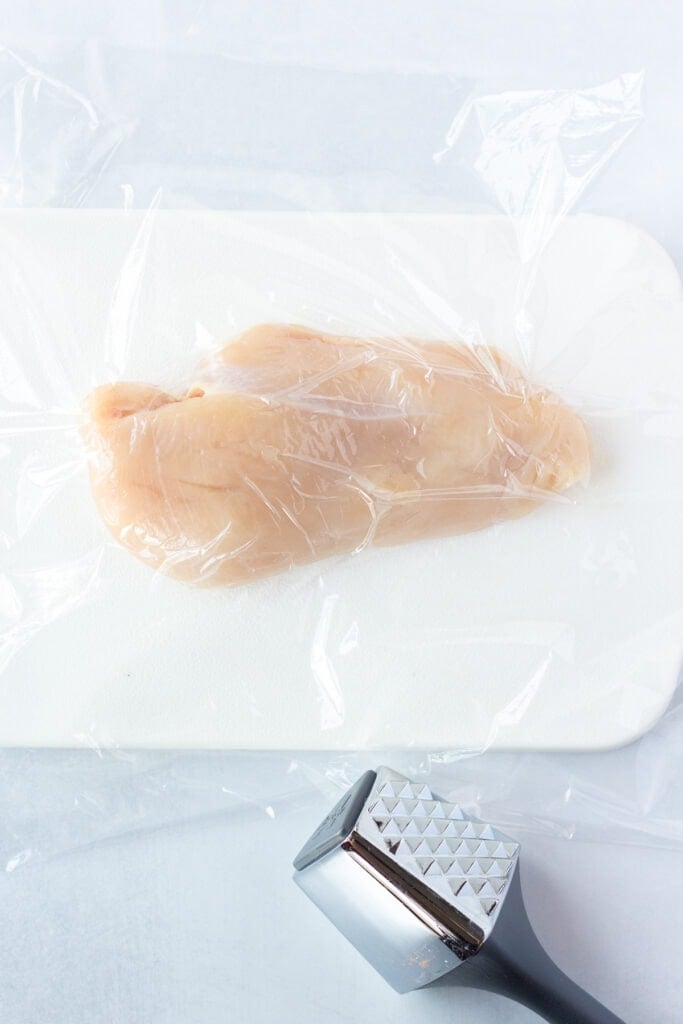 A raw chicken breast on a white cutting board with plastic wrap over it.