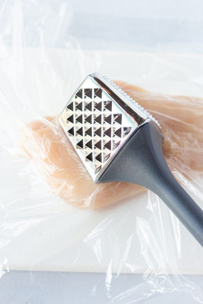 A meat mallet pressing into a raw chicken breast with plastic wrap over it.