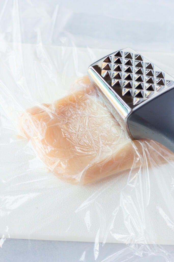 A meat mallet coming up from striking a raw chicken breast with plastic wrap coming over it.