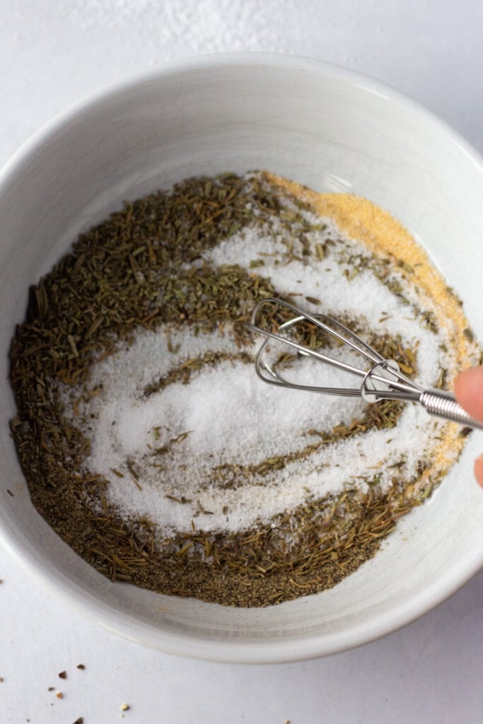 A small whisk stirring together kosher salt, herbs, and spices in a small white bowl.