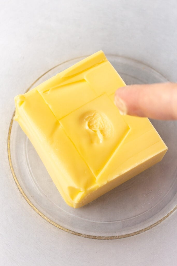 Close up of a slab of butter in a large clear bowl with a finger that has butter on it having just touched it and left a sticky impression in the middle.