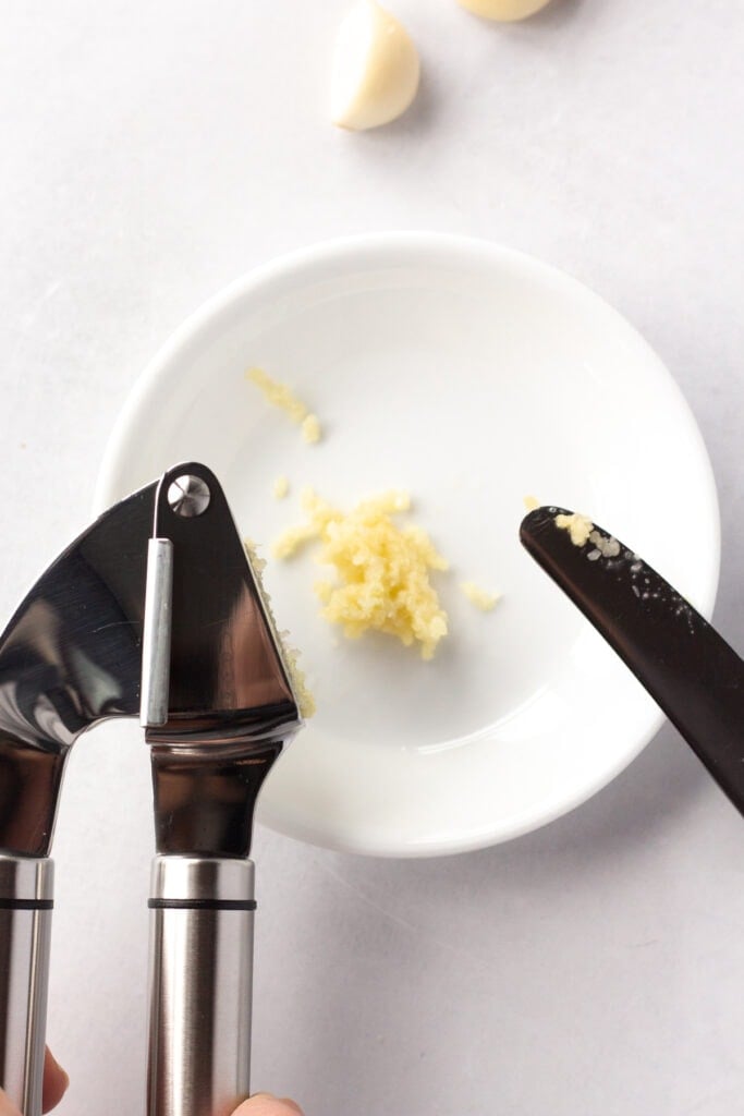 A garlic press pressing fresh garlic onto a small white plate with a knife on the side.