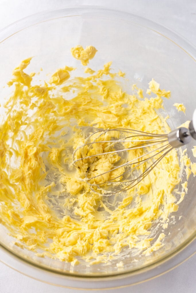 Butter being whipped with a handheld beater in a large clear bowl on a white background.