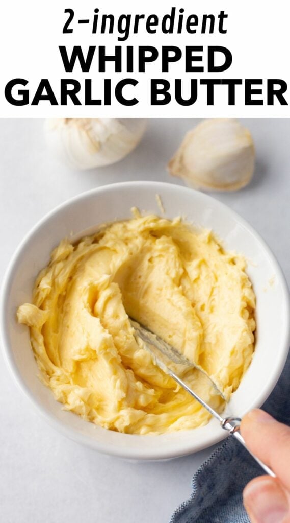 pin for 2 ingredient whipped garlic butter, with those words in black on a white background at the very top of an image of a small white bowl with whipped garlic butter in it. The butter has a small butter knife being pressed into it with a hand and fresh garlic cloves are near the bowl.