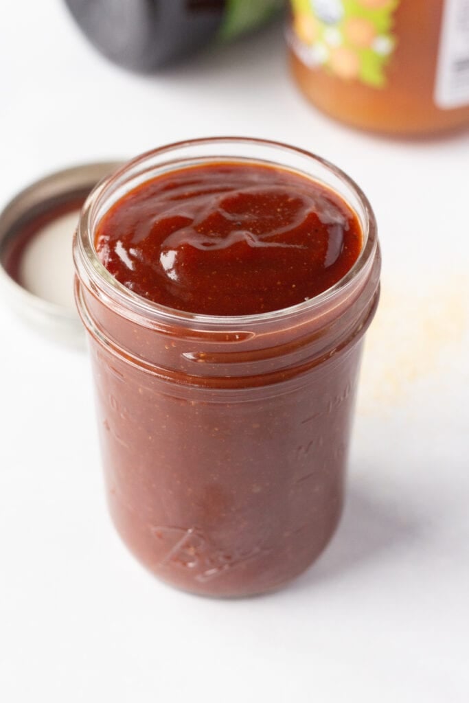 Close up of a small mason jar with apricot barbecue sauce in it on a white surface. In the background are jars with jam and molasses in them.