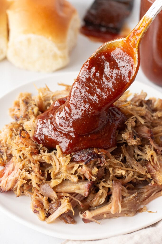 Close up of apricot barbeque sauce being poured over a white dish with pulled pork on it with a spoon.