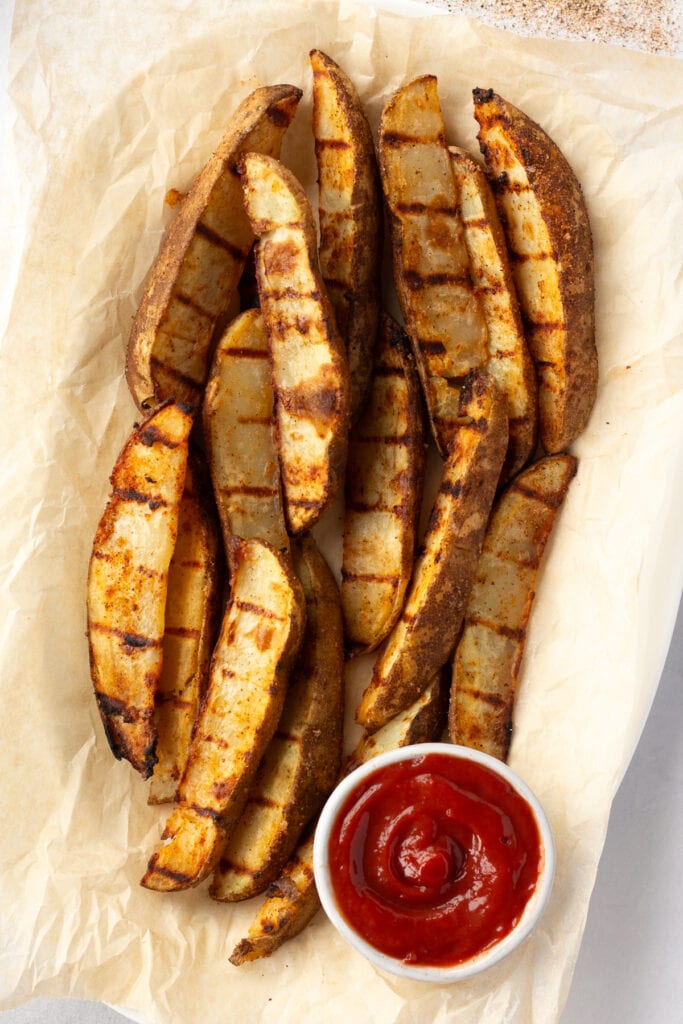top down shot of grilled potato wedges on top of brown parchment paper with a small white bowl of ketchup in the lower part of the frame.