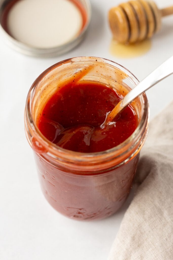 Close up angle shot of a small mason jar with bbq sauce in it. The jar has a spoon jutting out of it. Behind the jar are a lid and a honey stirrer and a tan cloth napkin is to the right of the jar.