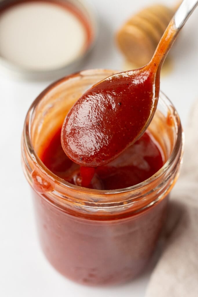 Close up of a spoon dipping into a small mason jar with a honey bbq sauce in it. The mason jar lid and a honey stirrer are in the background and everything is on a white surface.
