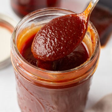 a small mason jar filled with maple bbq sauce that has a spoon bringing sauce out of it. The jar is on a white surface and in the background is a lid, a small jar of maple syrup, and a brush with bbq sauce on it.