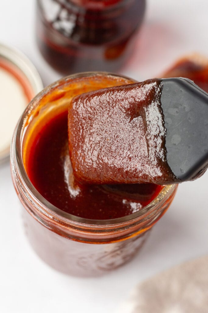 Close up of a basting brush being dipped into a small mason jar of bbq sauce that's resting on a white surface. In the background is a lid, a small container of maple syrup, a tan cloth napkin, and a spoon with bbq sauce on it.