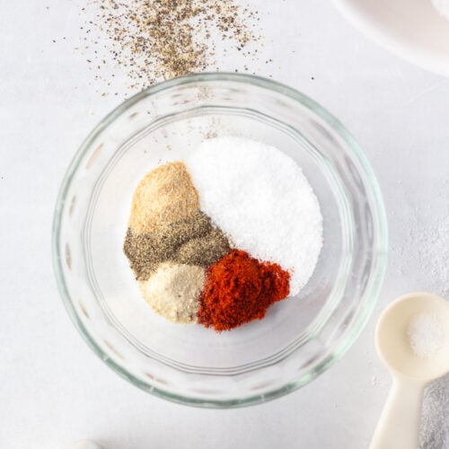 Top down shot of a small clear bowl with spices in it, including kosher salt, ground black pepper, garlic powder, onion powder, and smoked paprika. Measuring spoons and spice jars are off to the side of the image.