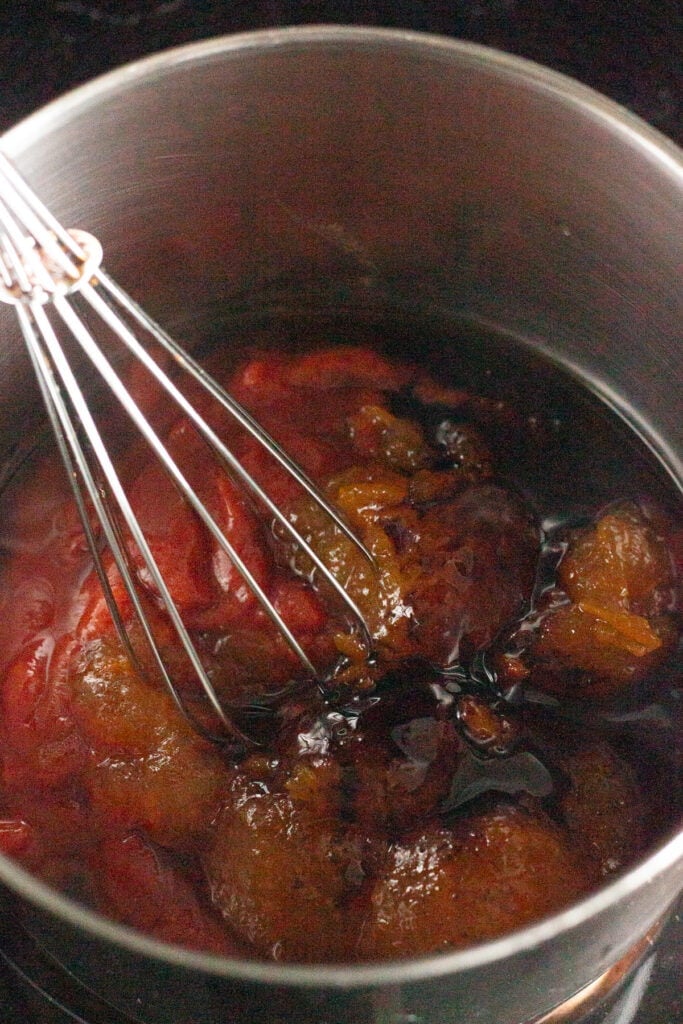 A close up of a pan with ingredients for apricot bbq sauce in it, with a whisk about to stir them together.