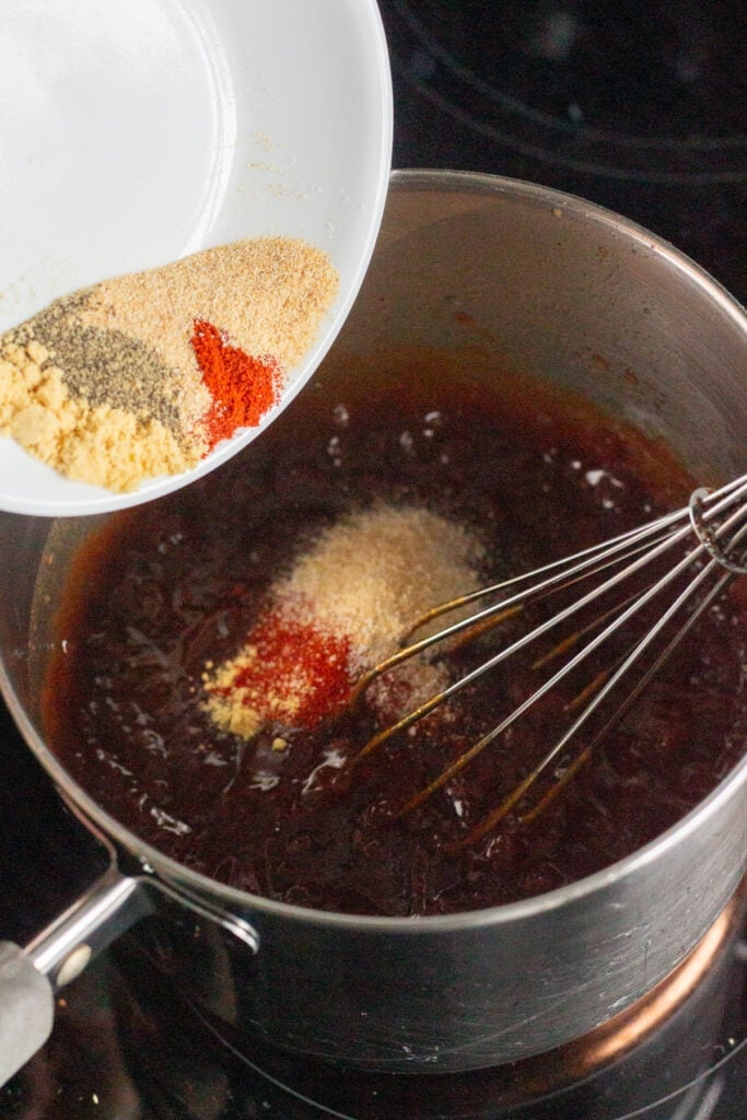 Close up of a pot with ingredients for apricot bbq sauce in it, with spices being poured out of a white plate into the pan. The pan also has a whisk in it.