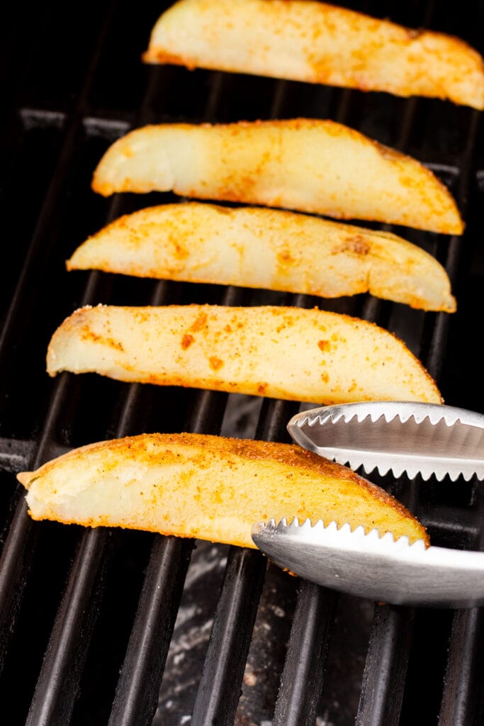 par cooked russet potato wedges being place on a hot grill with a pair of silver tongs.
