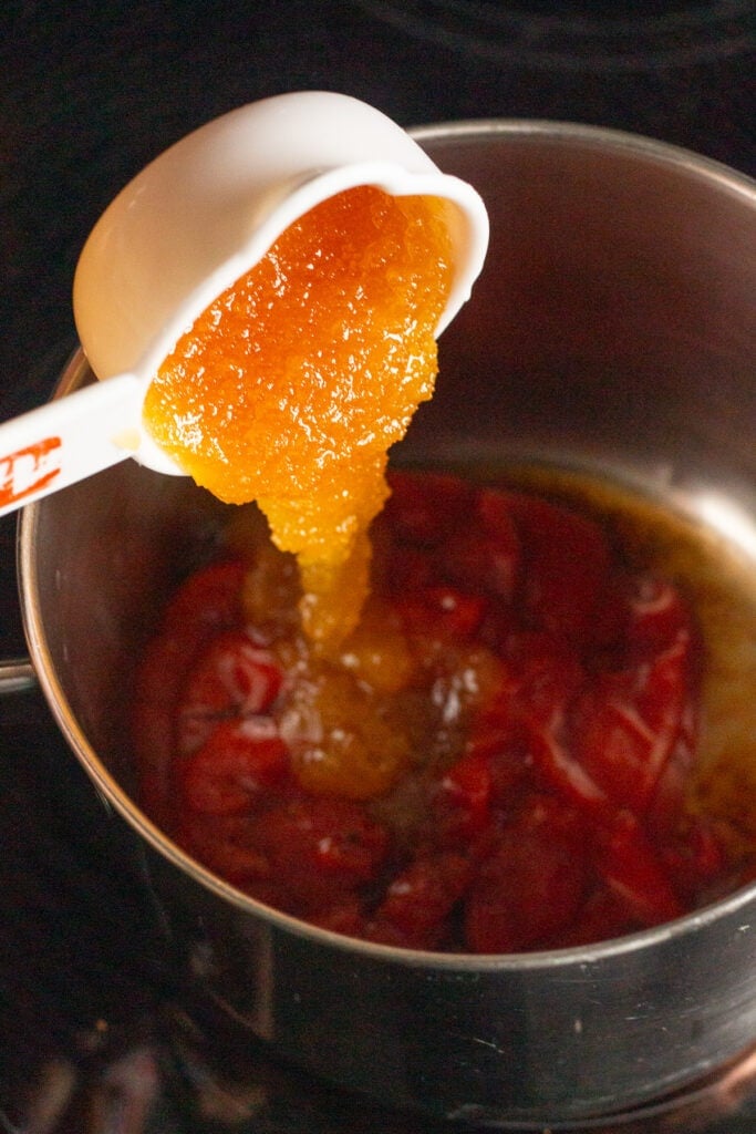 Honey being dumped out of a white measuring cup into a saucepan on the stove with ketchup and other ingredients in it.