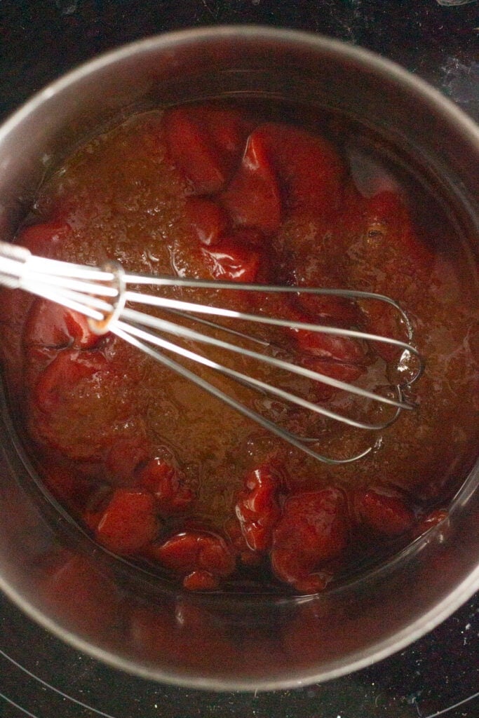 Top down shot of a whisk in a saucepan that's on the stove that also has honey, ketchup, and other ingredients in it.