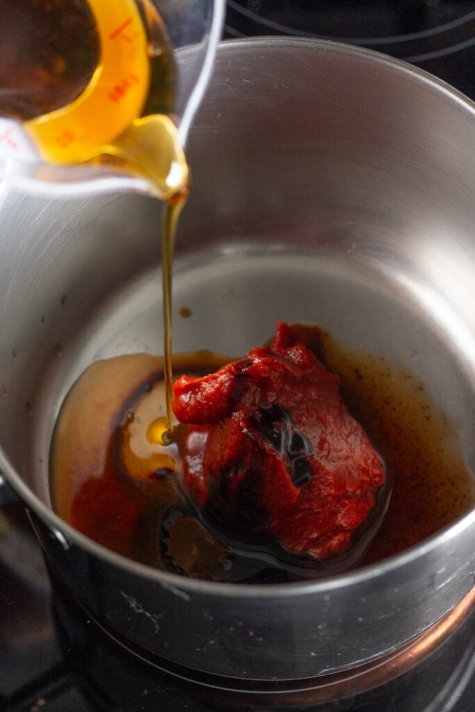 pouring maple syrup into a small saucepan on the stove that has tomato paste in it.