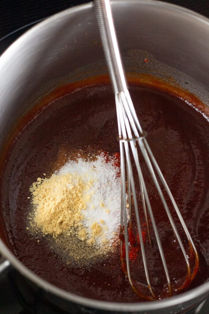 Close up of a sauce pan with a red tomato mixture in it with white, red, black, and yellow spices dumped into it. A whisk is also in the pan.