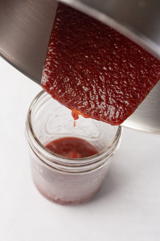 Close up of bbq sauce being poured out of a sauce pan and into a small mason jar on a white surface.