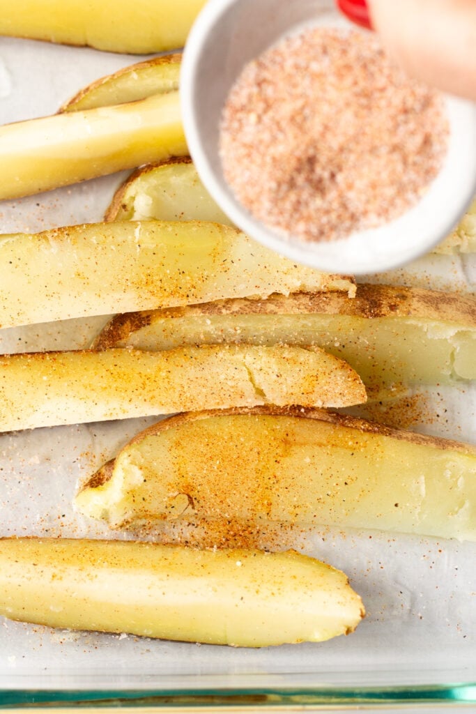 A seasoning mix being sprinkled out of a small white bowl and onto par-cooked potato wedges in a glass dish.