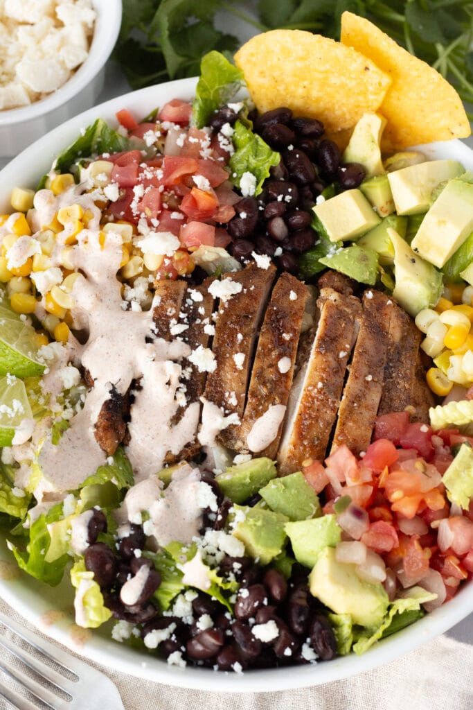 Close up of a chicken taco salad with strips of grilled chicken, lettuce, avocado, pico de gallo, black beans, corn kernels, tortilla chips, and a creamy taco dressing.