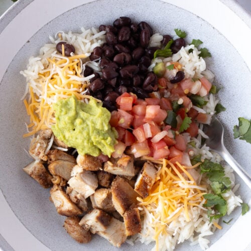 Top down shot of a gray bowl with white rice, chicken, pico de gallo, guacamole, black beans, and cilantro in it.