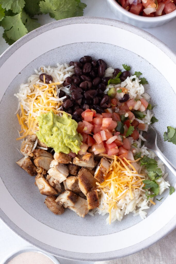 Top down shot of a gray bowl with white rice, chicken, pico de gallo, guacamole, black beans, and cilantro in it.