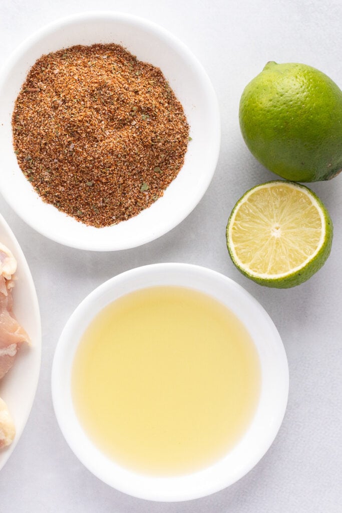 Top down shot of a ingredients for taco marinade, including a white plate with oil, a white plate with spices, and one and a half limes, all on a white surface.