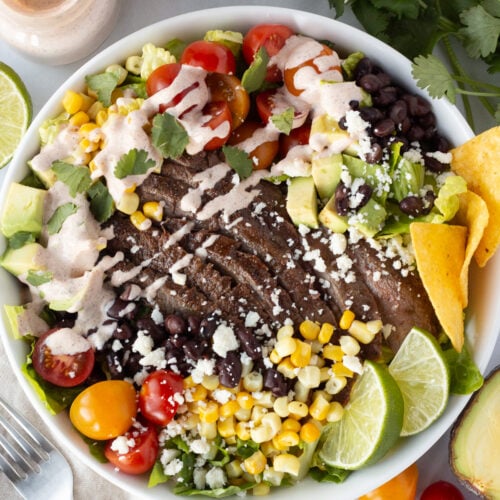 Top down shot of low white bowl filled with a steak taco salad, including lettuce, red and orange cherry tomatoes, corn kernels, black beans, tortilla chips, lime wedges, cilantro, and a dressing drizzled over it. Ingredients for the salad, as well as a tan cloth napkin, surround the bowl.