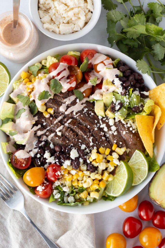 Top down shot of low white bowl filled with a steak taco salad, including lettuce, red and orange cherry tomatoes, corn kernels, black beans, tortilla chips, lime wedges, cilantro, and a dressing drizzled over it. Ingredients for the salad, as well as a tan cloth napkin, surround the bowl. 