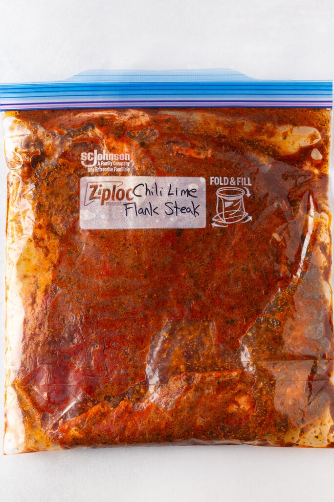 Top down shot of a raw flank steak in a red marinade in a gallon size sealable plastic bag. On the bag the words "chili lime flank steak" are written in black marker.
