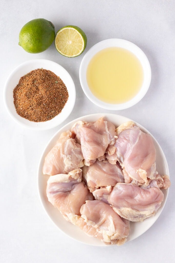 Top down shot of ingredients for chili lime chicken, including raw boneless skinless chicken thighs, one and a half limes, oil, and a seasoning blend.