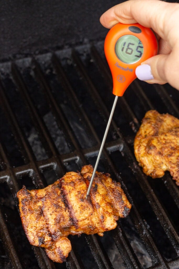 Close up of a hand measuring the internal temp of a boneless skinless chicken thigh on a gas grill. The meat thermometer is orange and reads 165F.