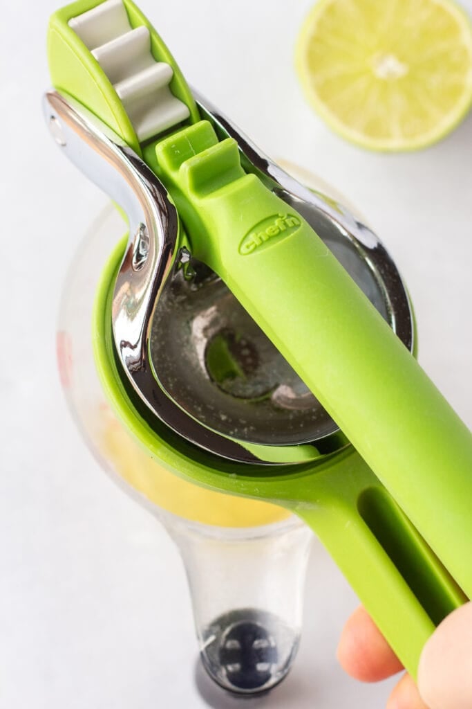 Close up of a lime being juiced into a measuring cup. The cup rests on an off-white surface and half a lime is in the background.