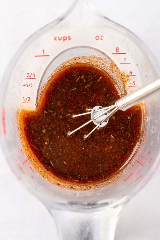 Top down close up of a chili lime marinade that's a dull red color in a plastic liquid measuring cup with a small whisk sticking out on the right side.
