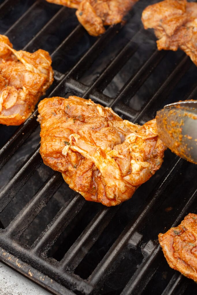 A pair of tongs placing marinated chili lime chicken thighs onto a gas grill.