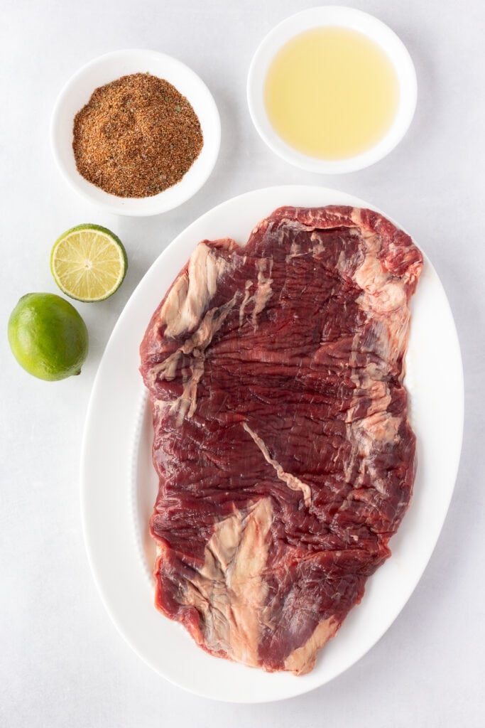 Top down shot of ingredients for chili lime flank steak, including a spice blend, limes, oil, and a raw flank steak on a white platter.