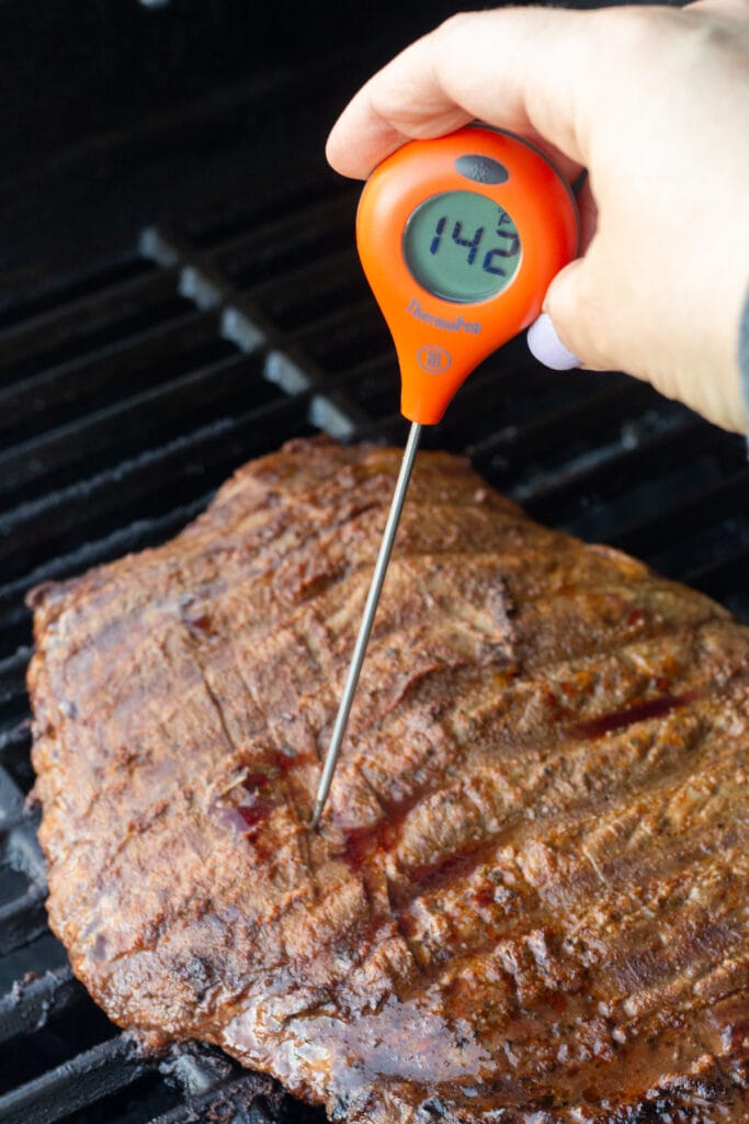 a hand placing a meat thermometer into a grilled flank steak, with the thermometer reading 142 degrees F.