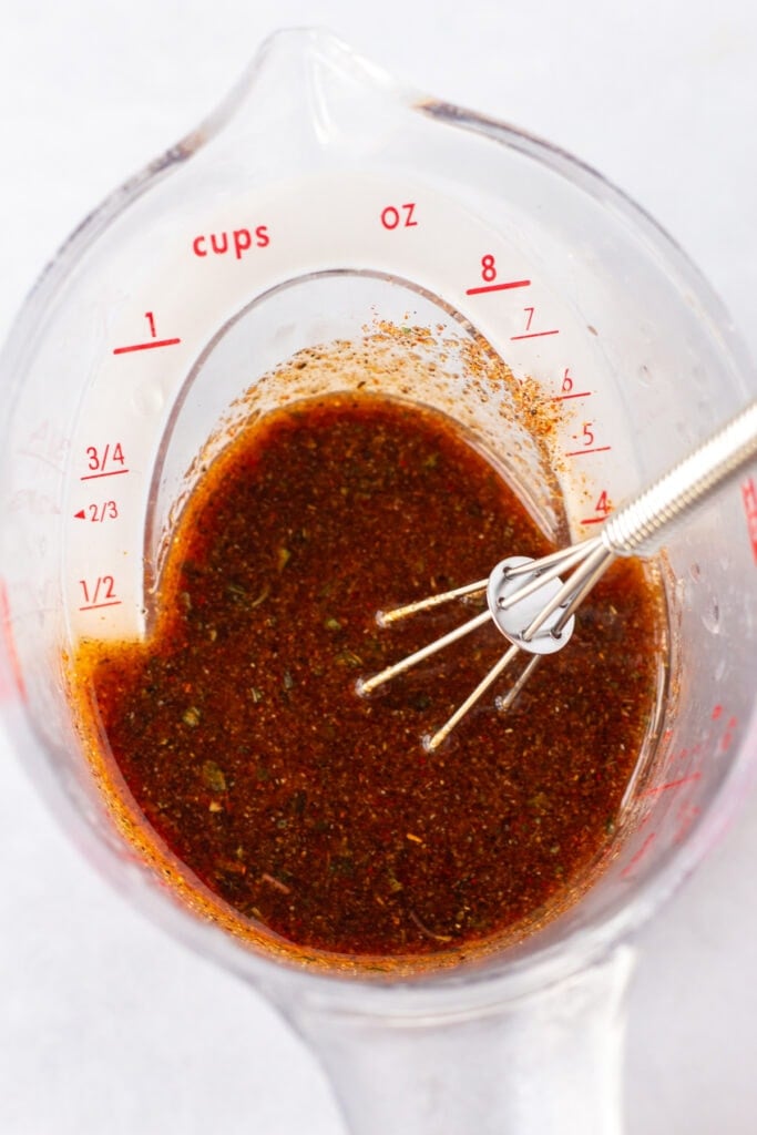 Close up of a liquid measuring cup with a red, chili lime marinade in it and a small whisk sticking out.