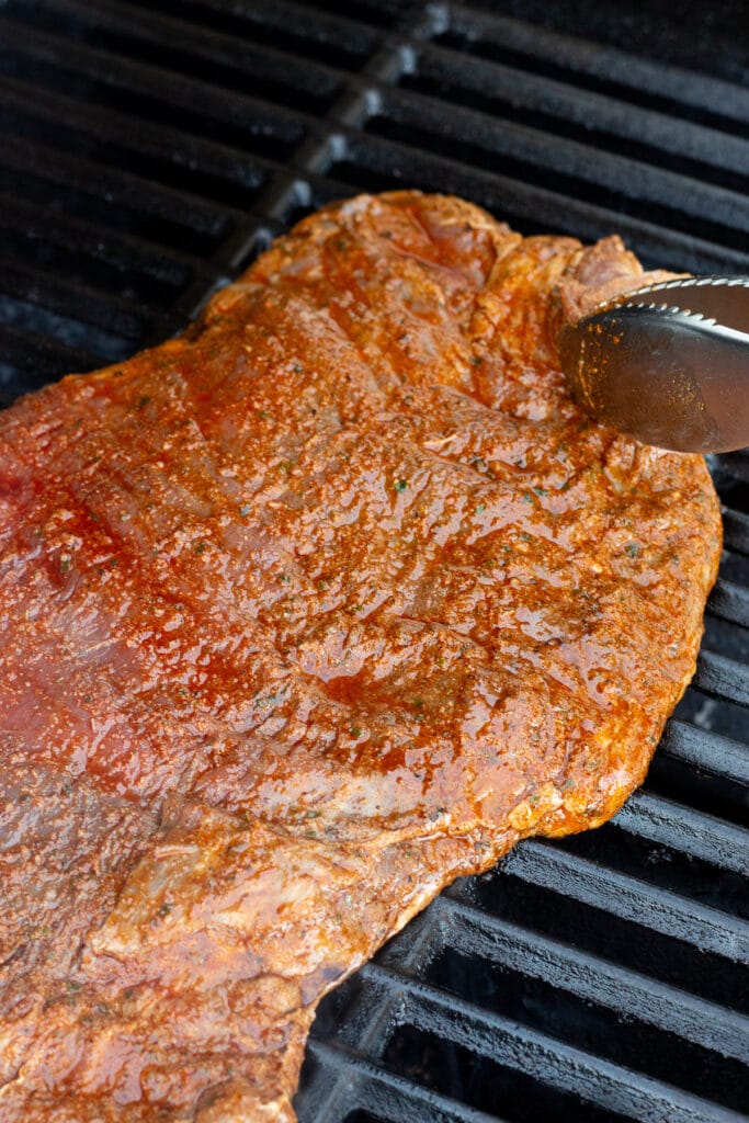 Placing a raw marinated flank steak on a hot grill with grilling tongs.