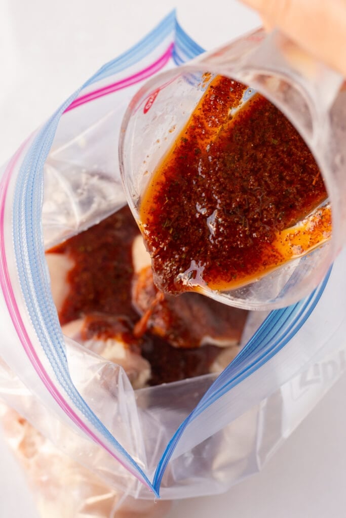 Close up of a red marinade being poured into a gallon sized ziploc bag with raw boneless skinless chicken thighs in it.